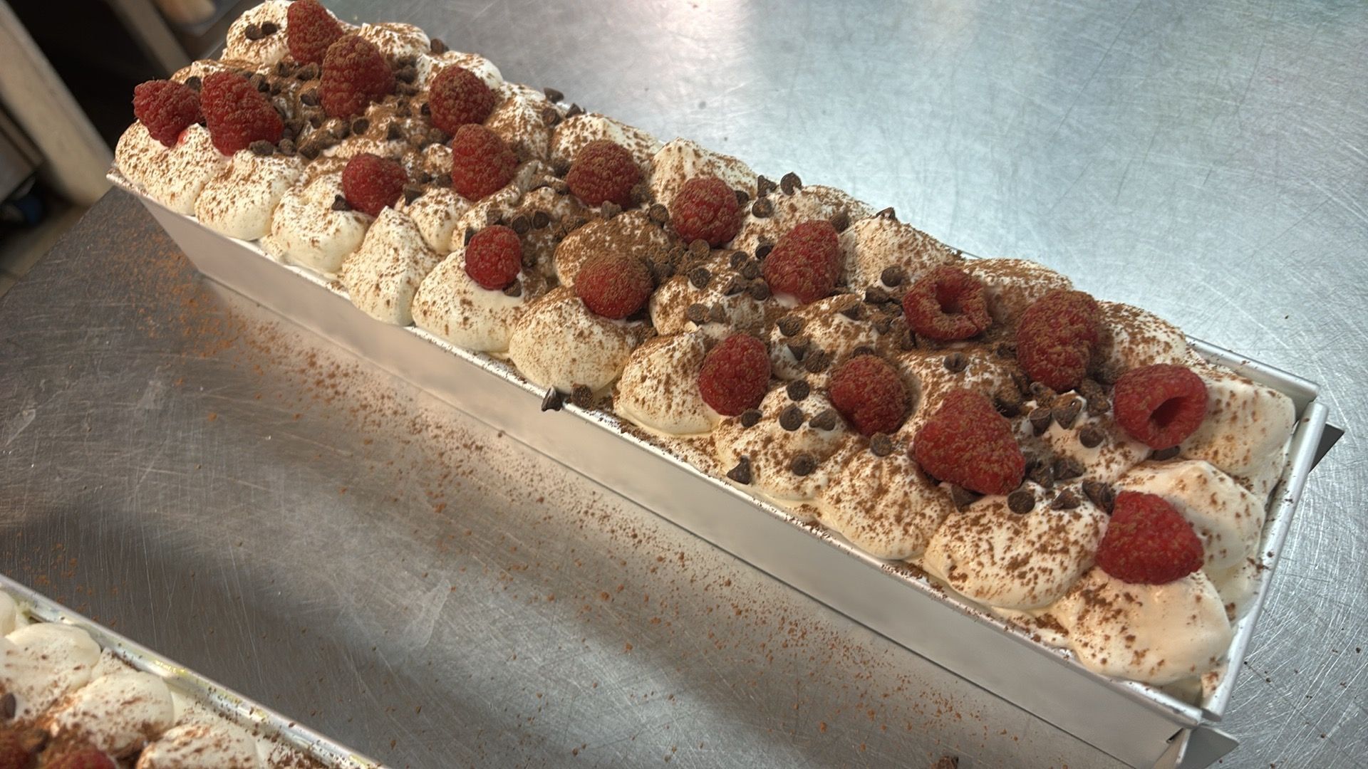 A rectangular tray of white frosted dessert topped with raspberries and dark chocolate shavings.