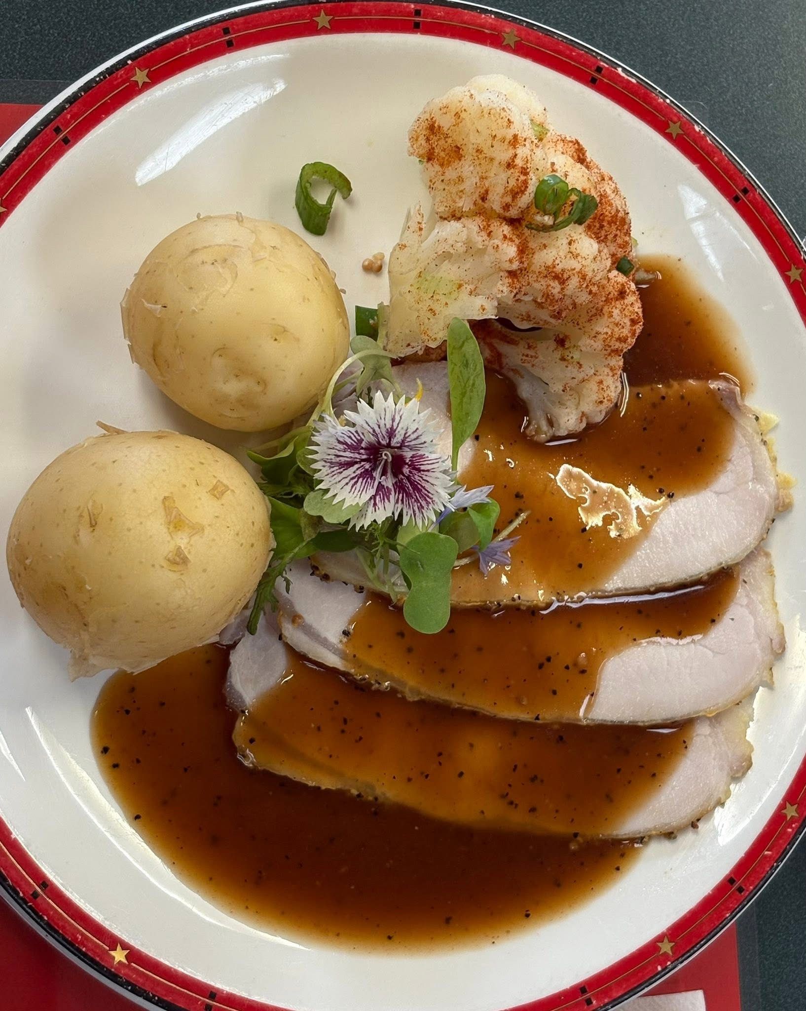 Three slices of roasted meat with brown gravy, two round boiled potatoes, and roasted cauliflower on a festive plate.