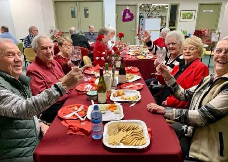 A group of people sitting at tables with red tablecloths, sharing wine and food during a celebration in a communal room.