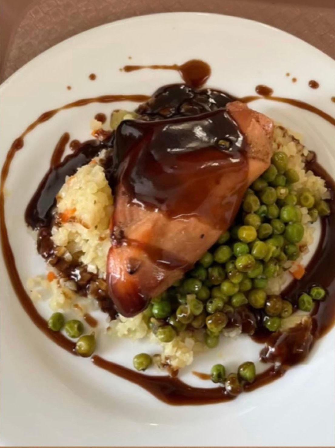 A plated meal featuring a piece of glazed poultry, a serving of rice, and a pile of green peas with a dark sauce drizzle.