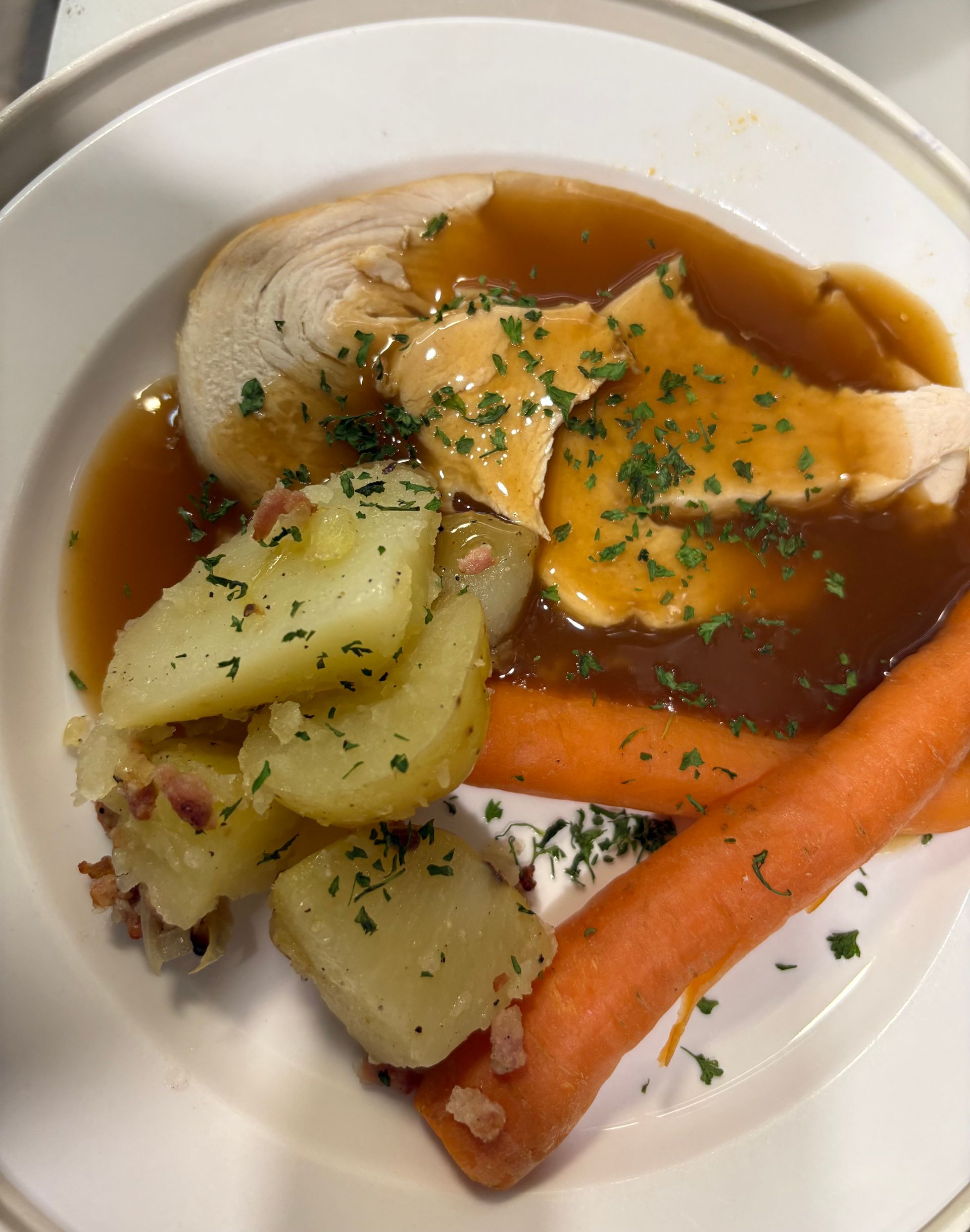A plated roast dinner featuring sliced meat in gravy, potatoes with bits of bacon, and whole carrots, topped with herbs.