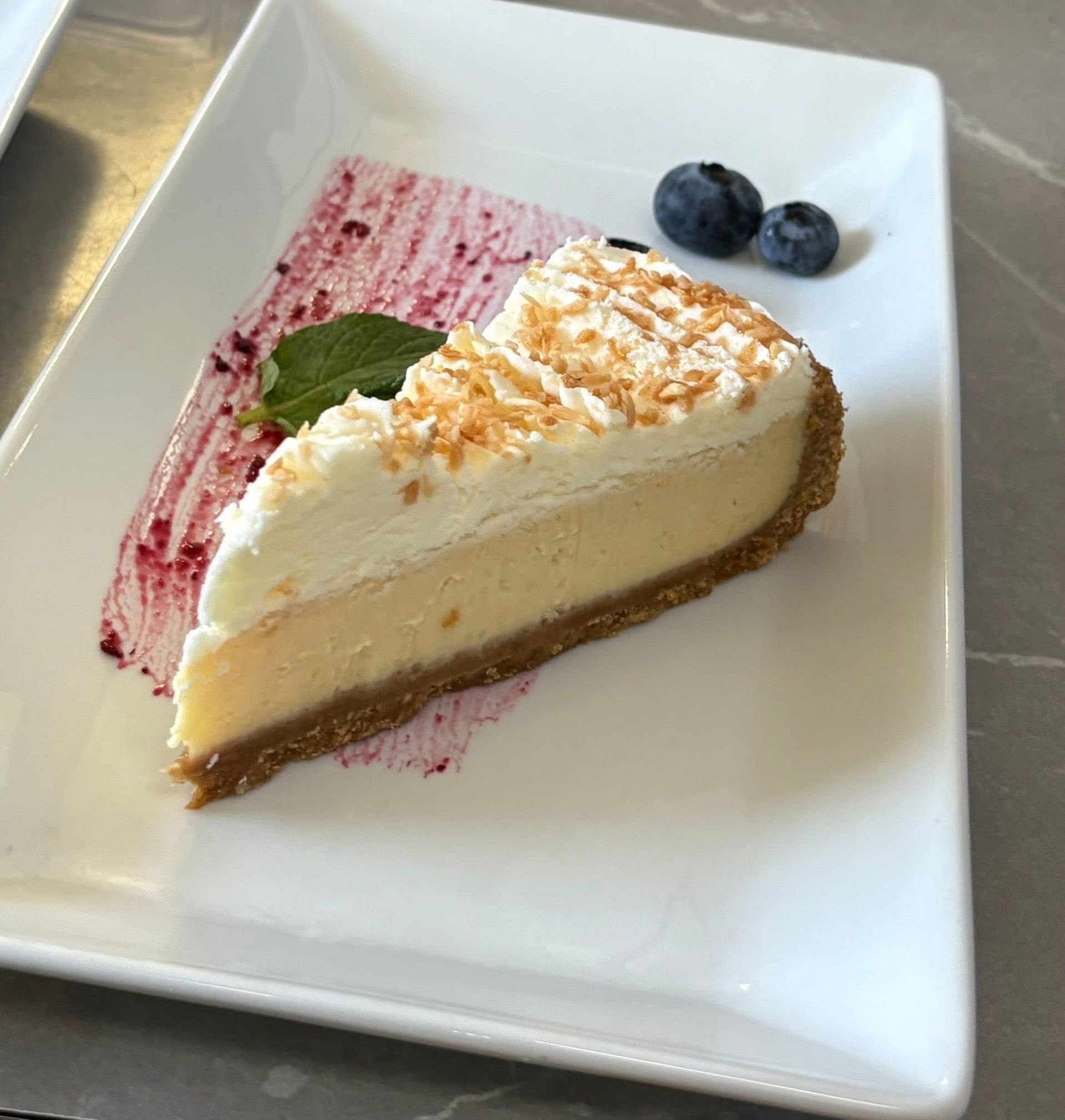 A slice of creamy coconut cheesecake topped with whipped cream and toasted coconut, served with berries and sauce.