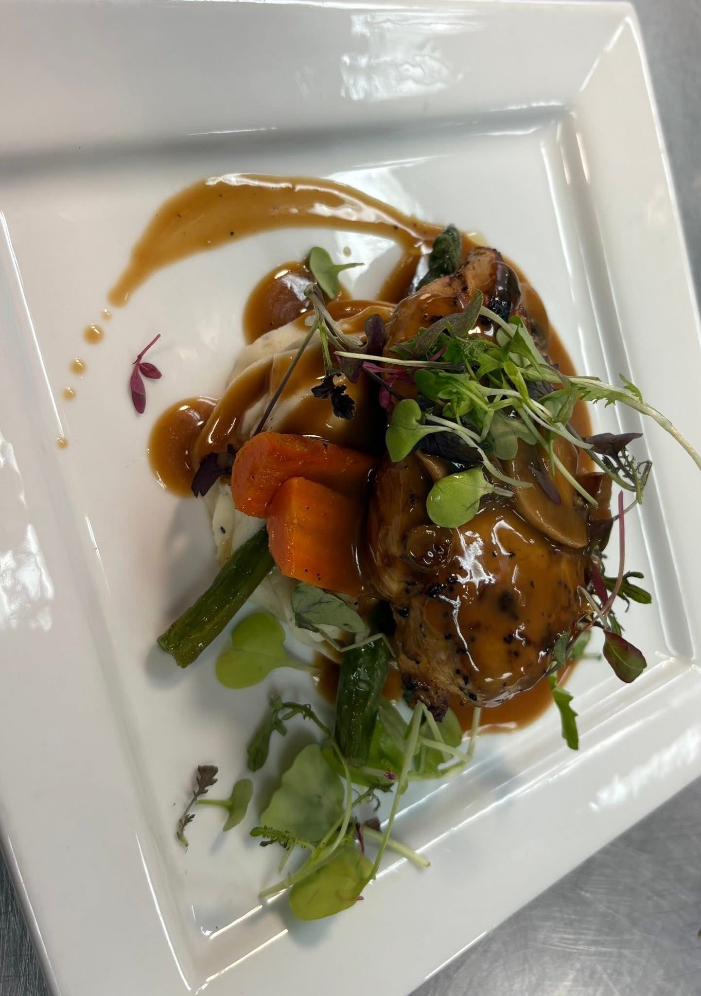 A plated dish featuring roasted chicken, carrots, and green beans topped with a brown mushroom sauce and microgreens.