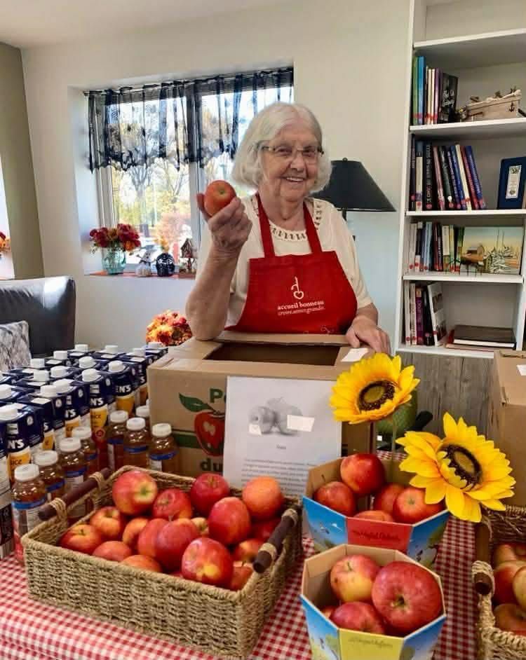 A person wearing a red apron stands behind boxes and baskets of apples, holding one up with a smile in a bright room.