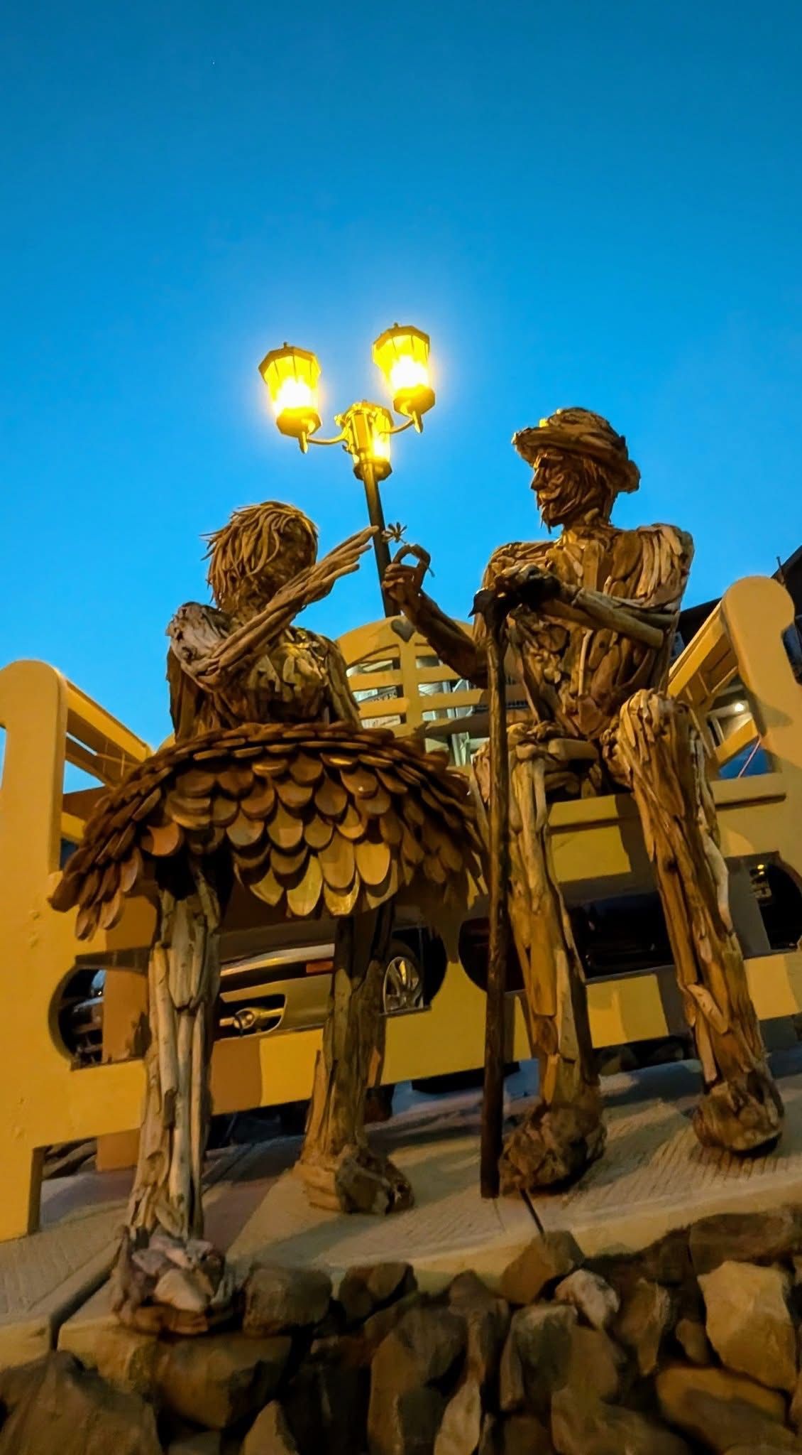 Two textured metal figures sit on a yellow bench, one holding an illuminated lamp post against a clear blue sky.