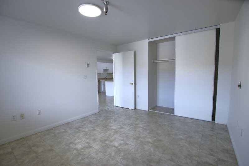 A bright, empty bedroom with white walls, tan tile flooring, a closet with sliding doors, and an open doorway.