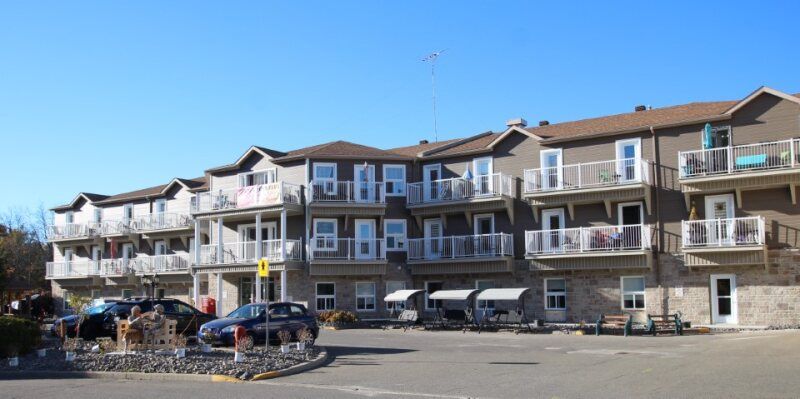 A multi-story brown residential building with balconies and a parking lot in front under a clear blue sky.