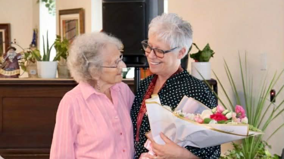 Two smiling individuals stand together, one in a pink shirt and one in a black-and-white top, holding a bouquet of flowers.
