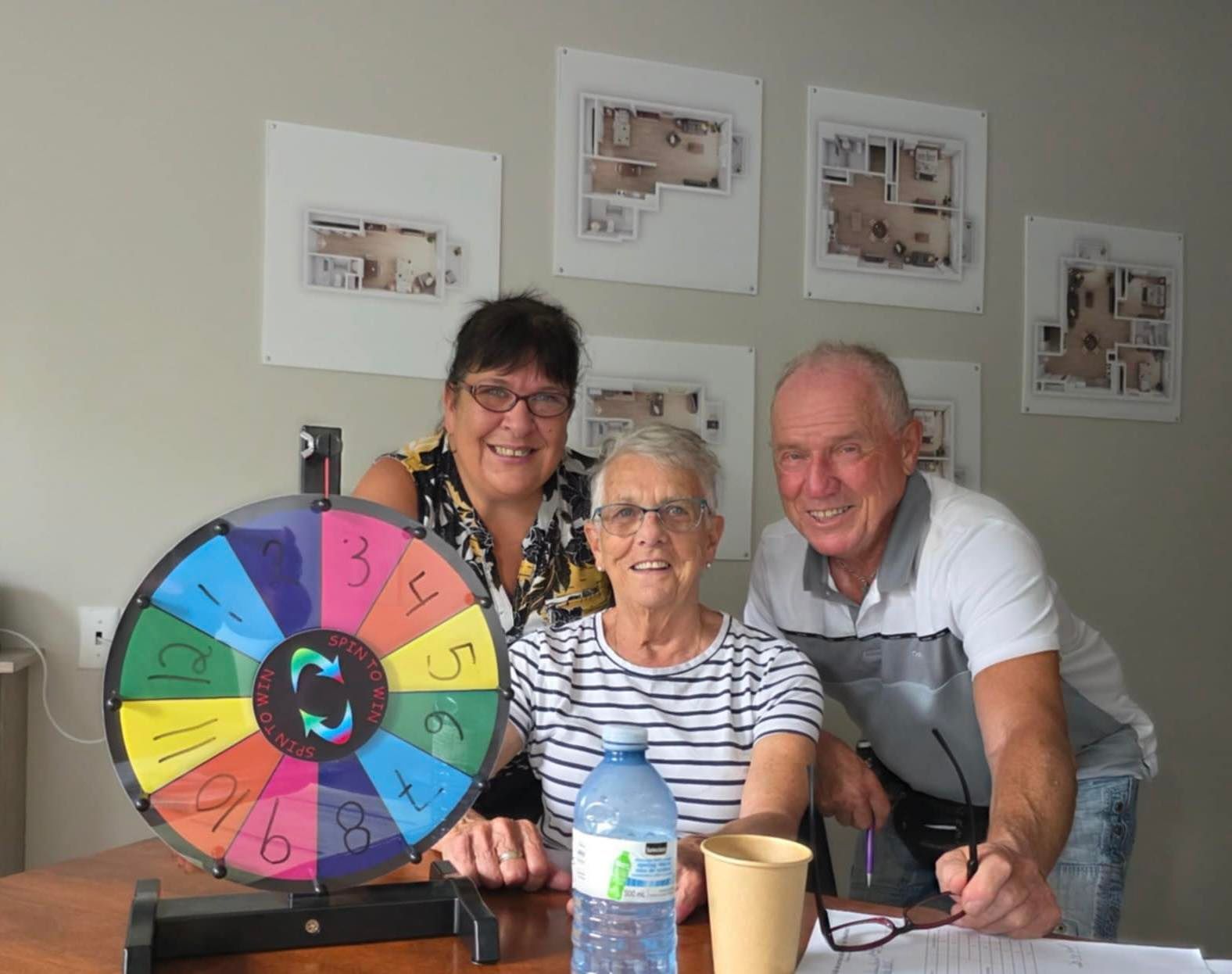 Three people smiling behind a table with a colorful numbered spinning prize wheel, water bottle, and papers.