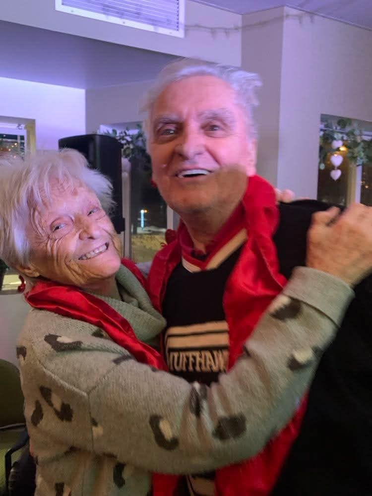 Deux personnes souriantes et enlacées, portant une écharpe rouge, dans une pièce baignée de lumière.