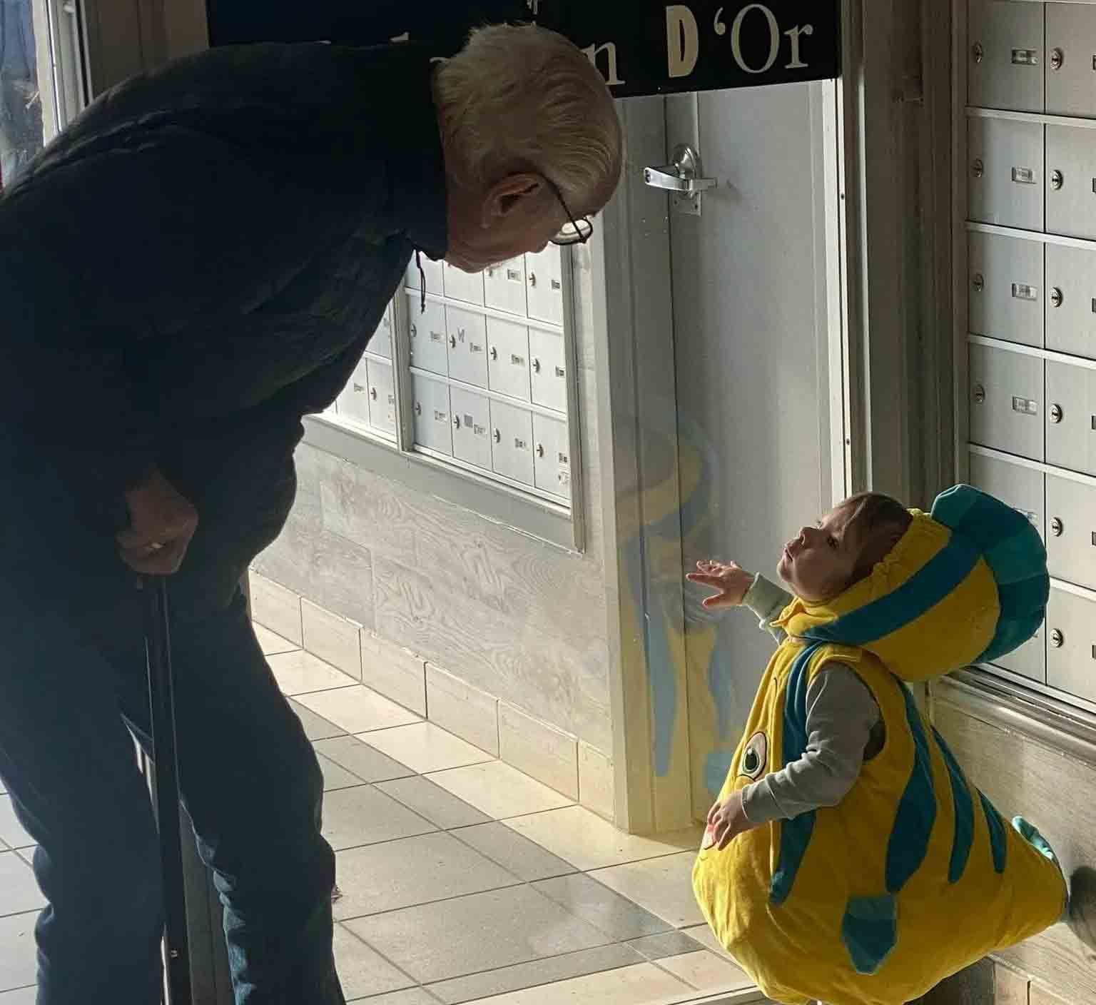 Dans une salle de courrier, une personne âgée s'appuyant sur une canne se penche vers un tout-petit déguisé en poisson jaune et turquoise.