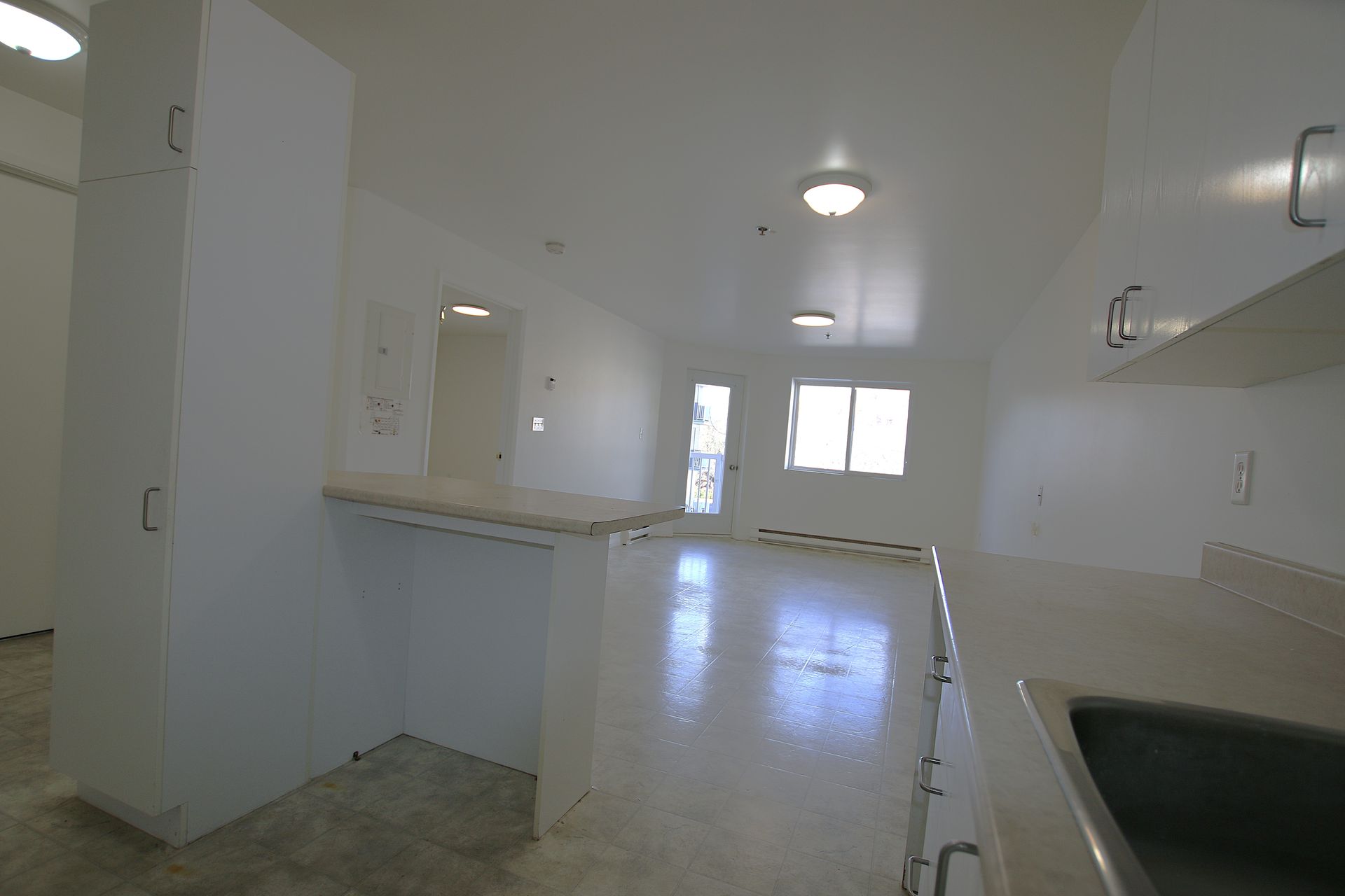 A bright, empty kitchen with white cabinets, a light countertop, and a living area with windows visible in the background.