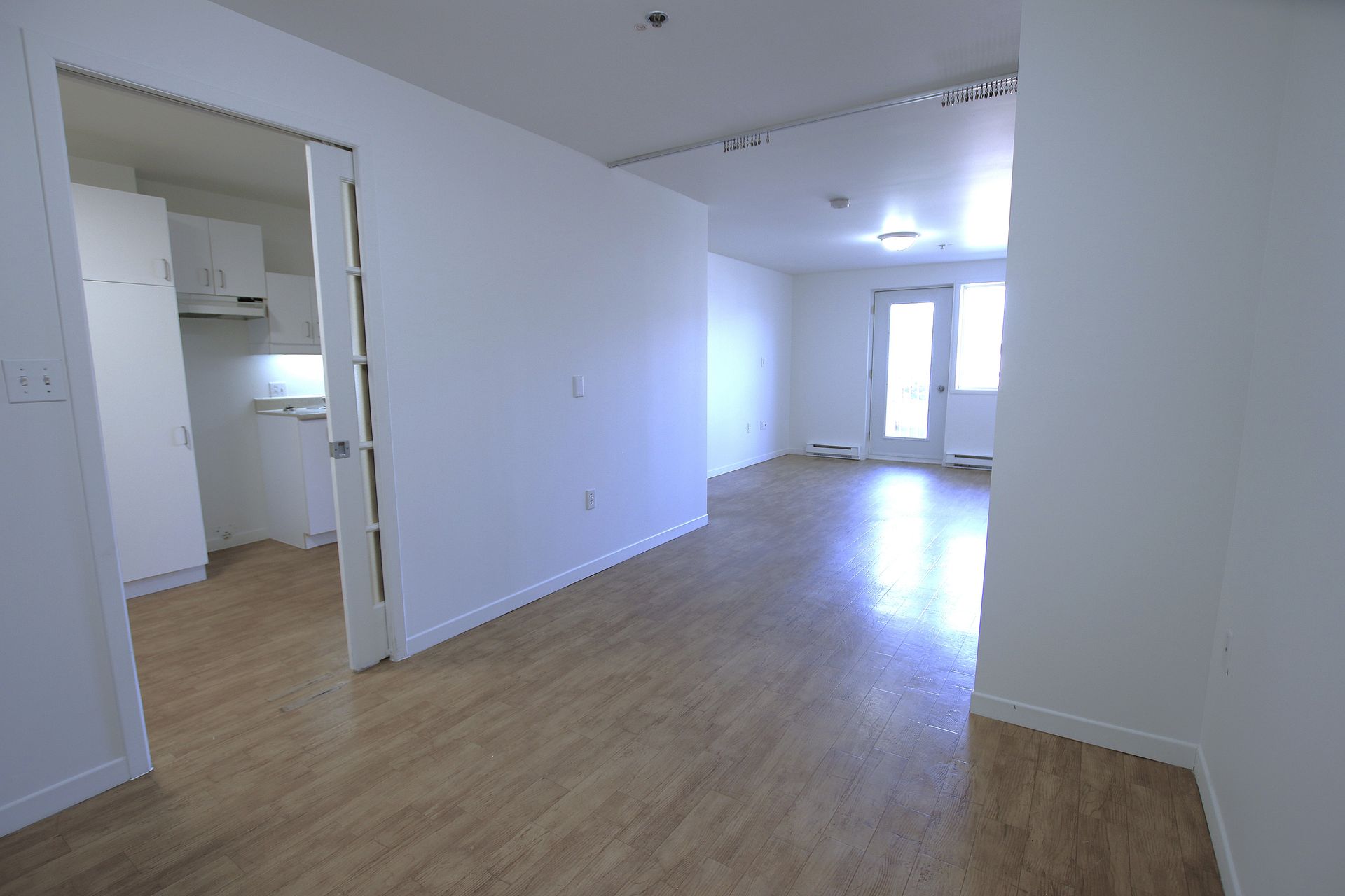 A bright, empty apartment interior with light wood-style flooring, white walls, and a doorway leading to a kitchen.