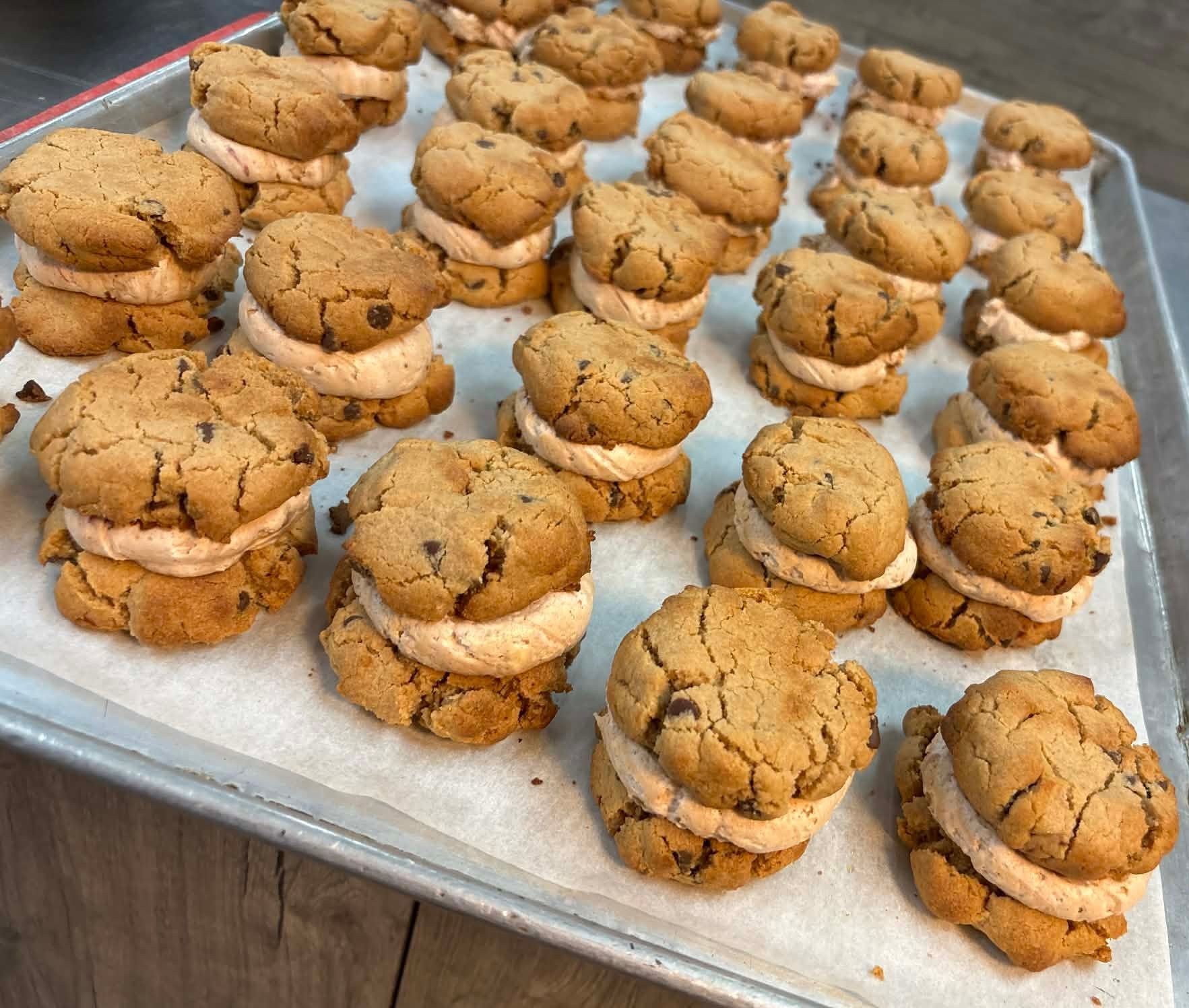 A baking tray filled with numerous cookie sandwiches featuring two chocolate chip cookies with a light orange filling.