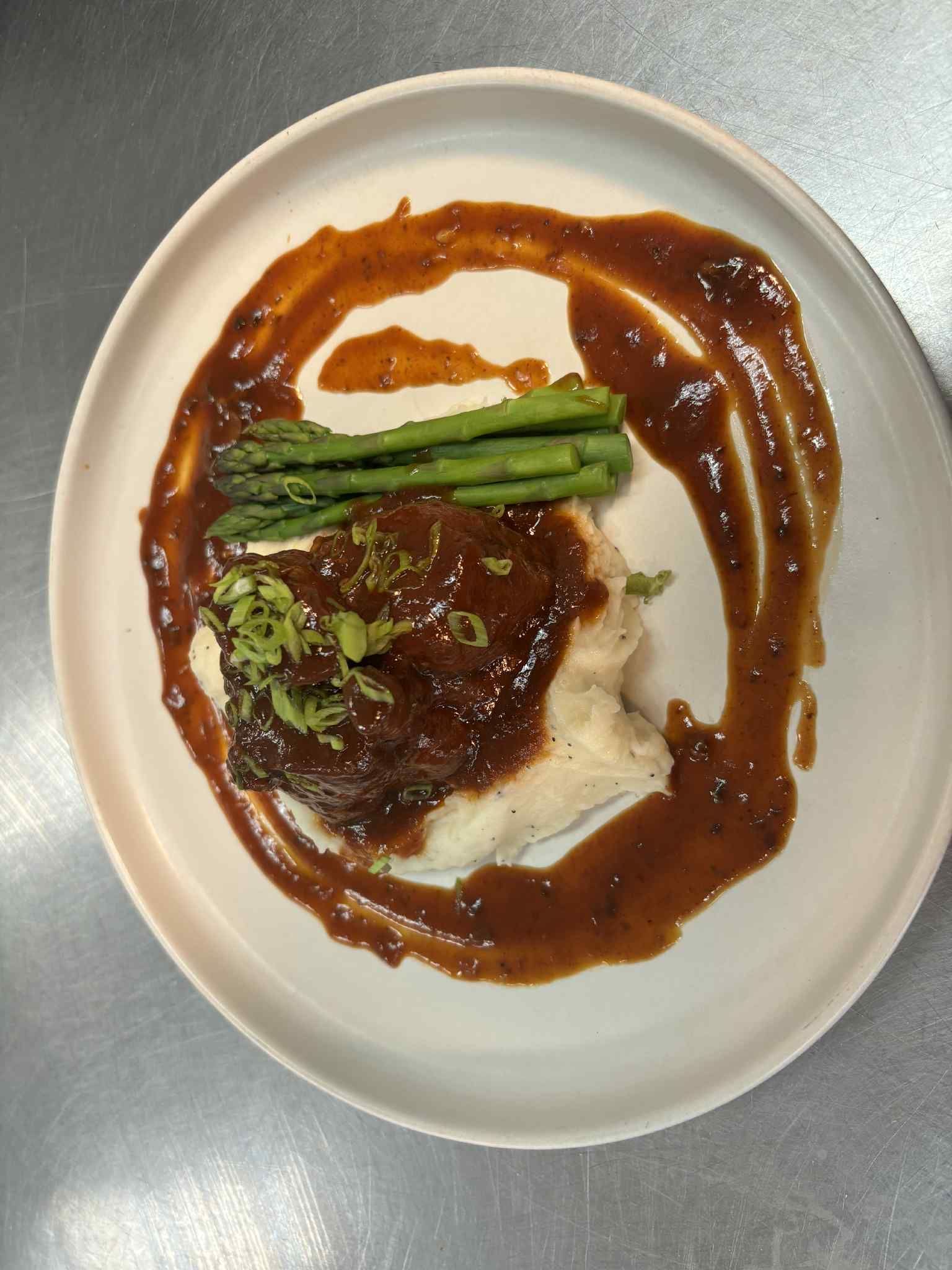 A plated meal featuring braised meat served over mashed potatoes with asparagus, topped with sauce and herbs.