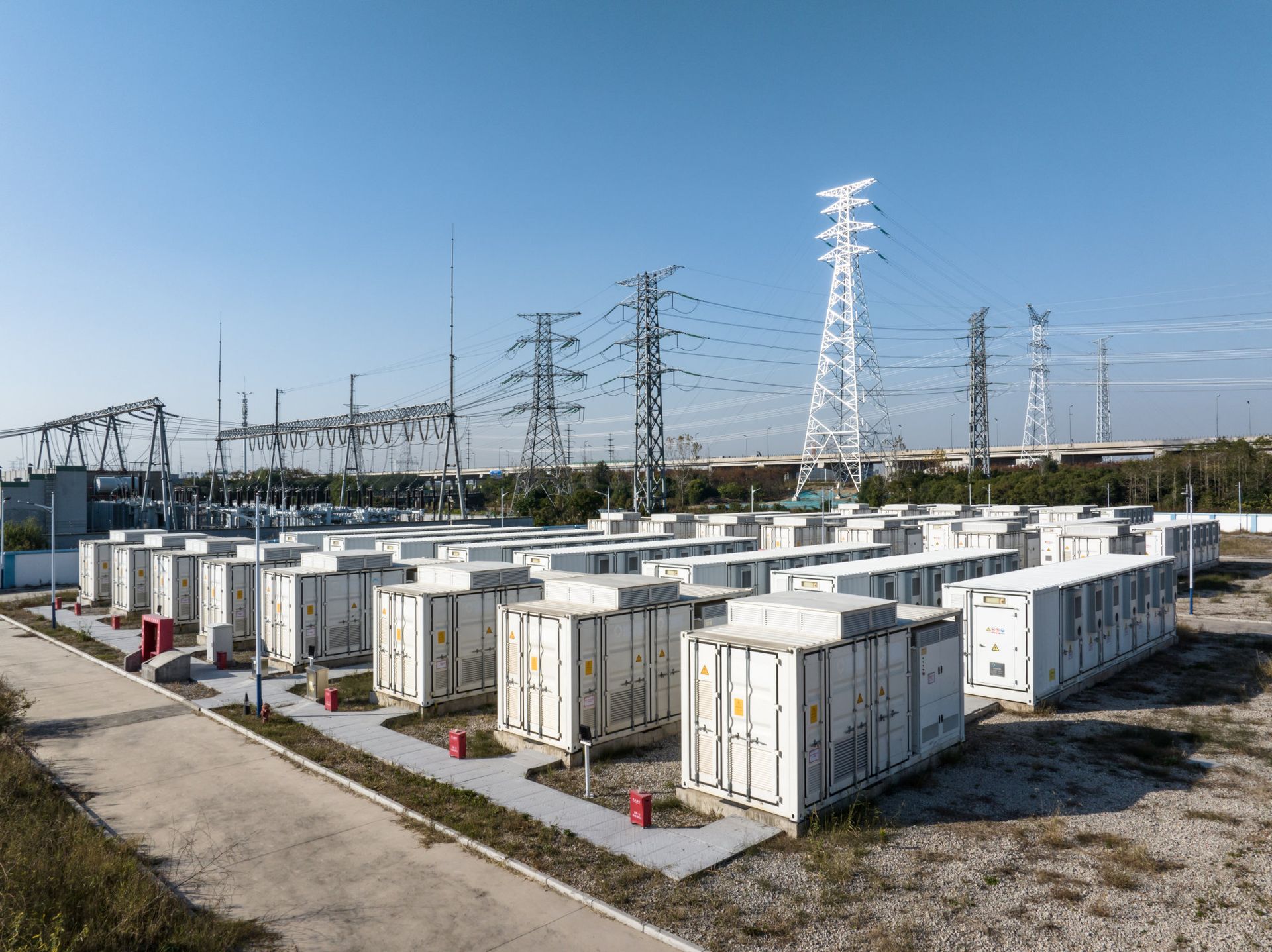 Rows of white electrical storage containers in a utility yard with power lines and transmission towers.