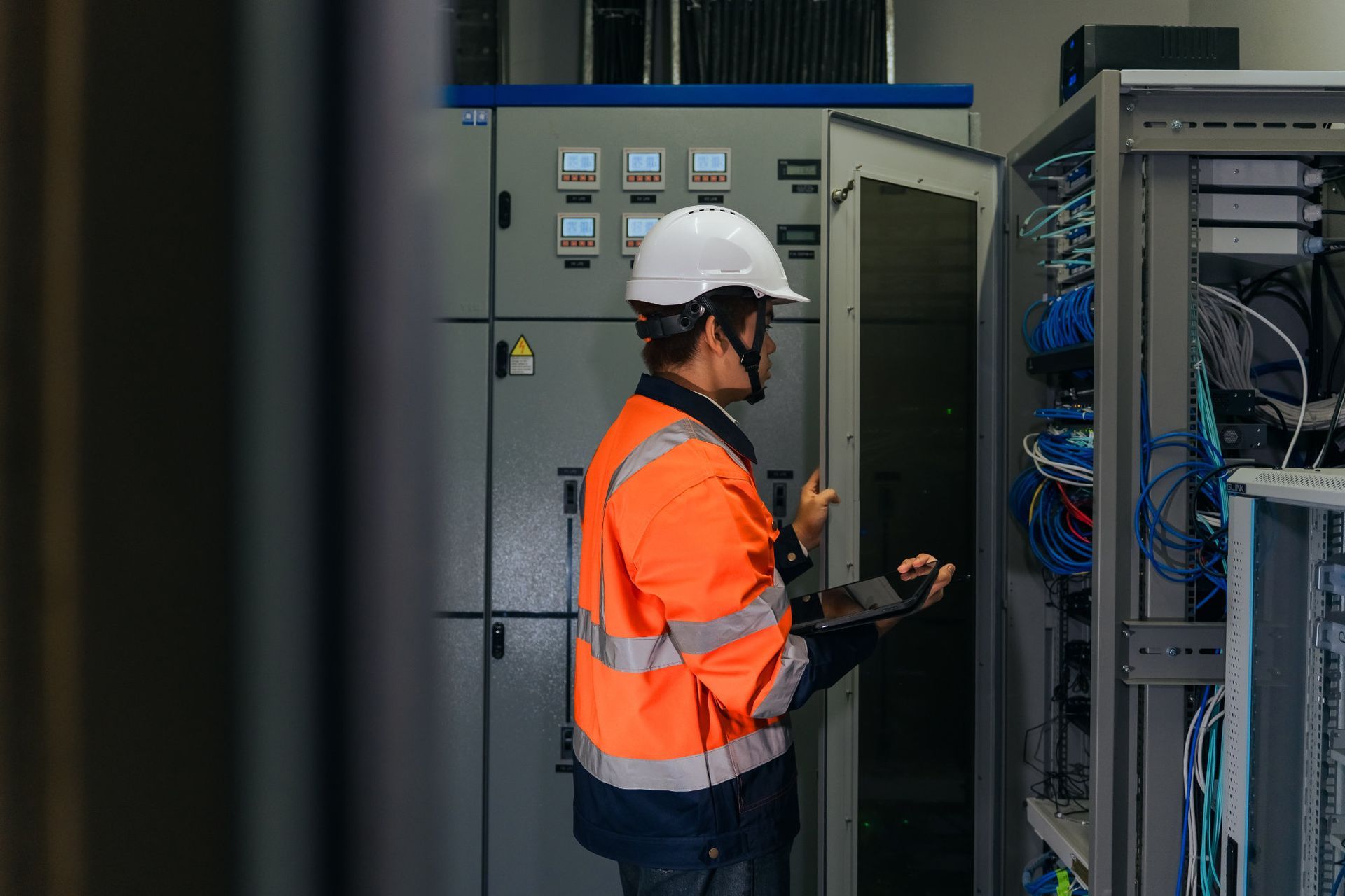 Technician in orange safety vest and hard hat inspecting electrical panels in a server room