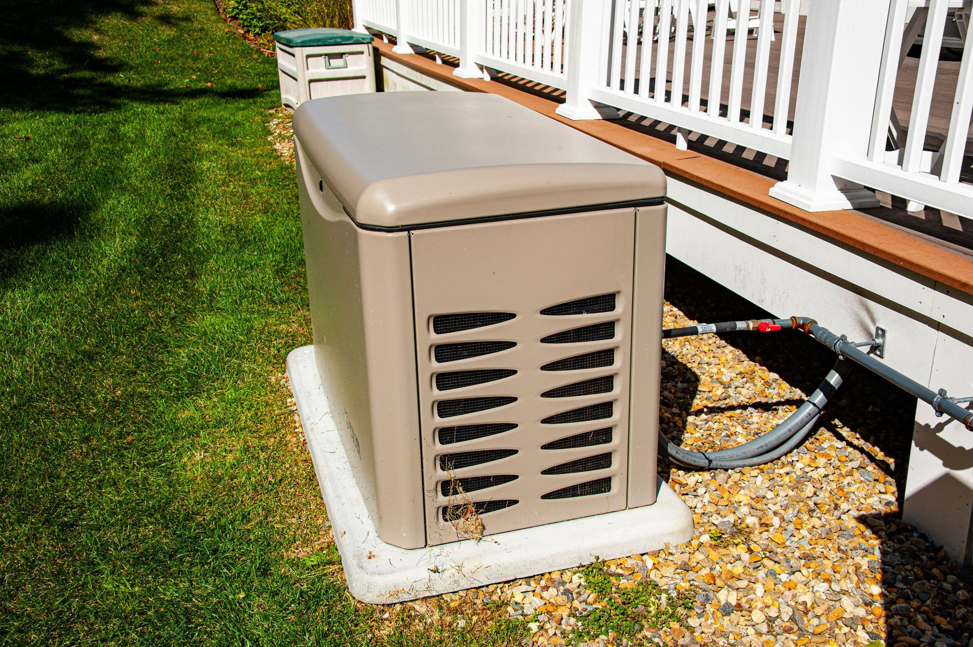 Outdoor beige HVAC unit beside a house deck, sitting on gravel next to a green lawn.