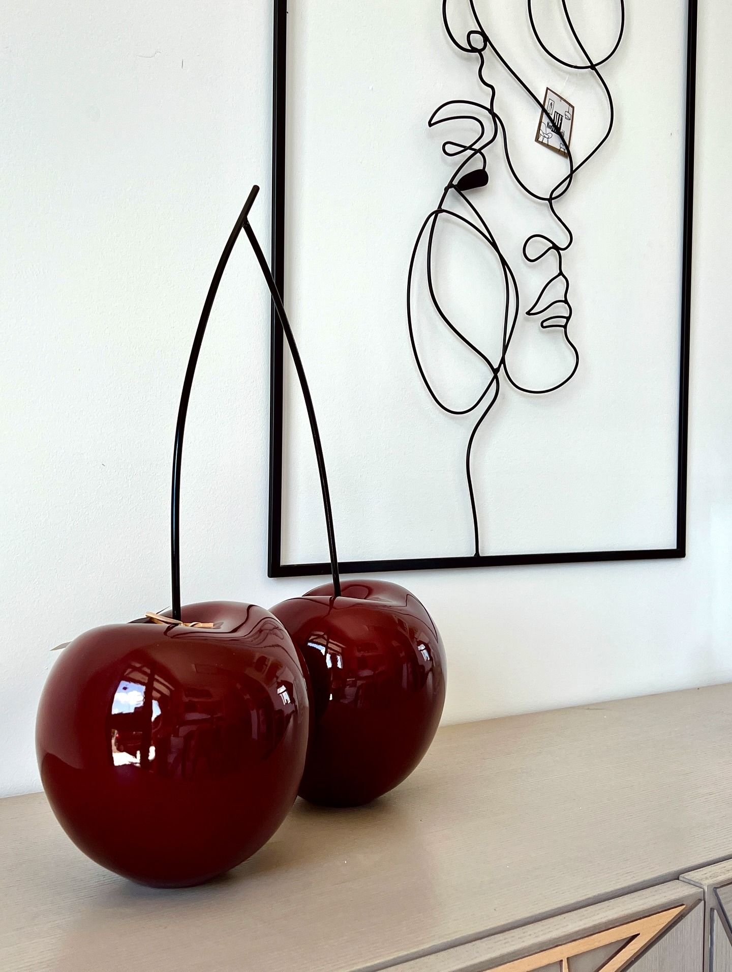 Due sculture di ciliegie rosse lucide su uno scaffale, con un volto in filo metallico sullo sfondo.