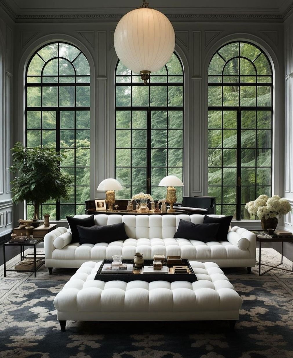 Elegante soggiorno con alte finestre ad arco che si affacciano sugli alberi, mobili bianchi e una grande lampada rotonda.