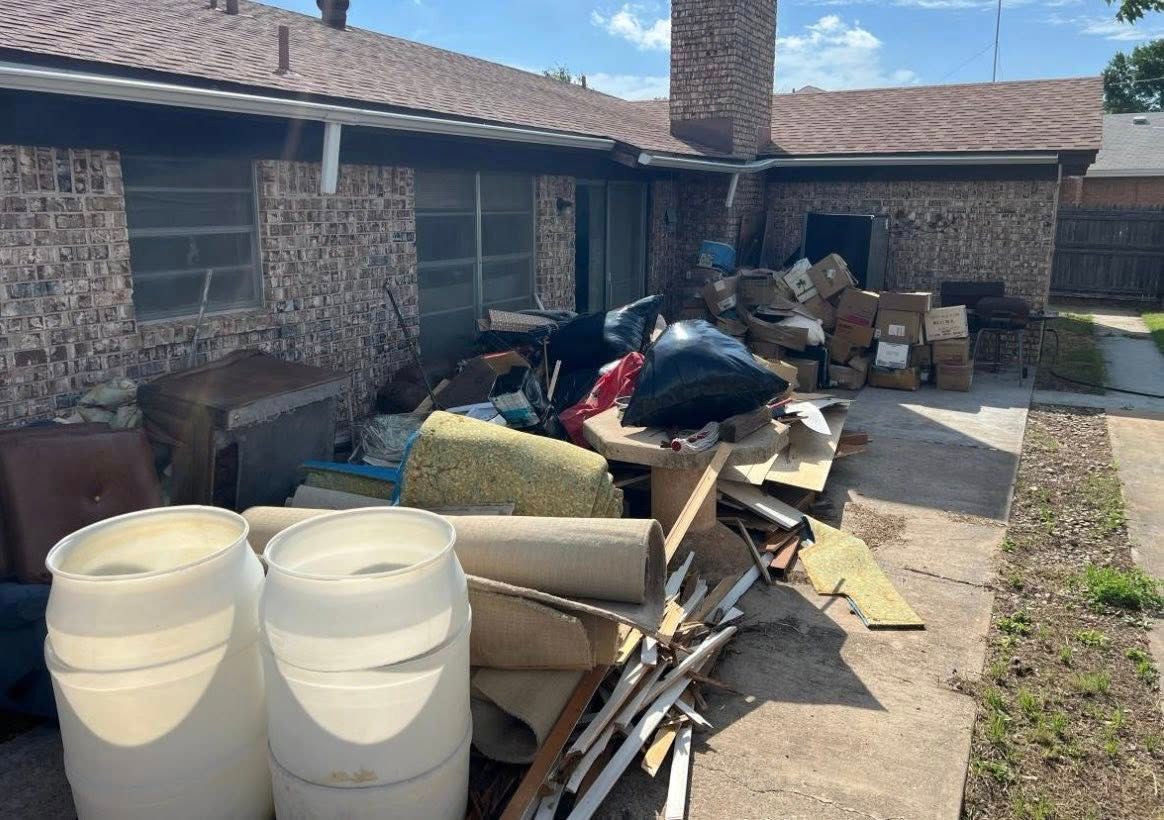 Piles of trash and bulky debris stacked outside a house along a walkway and brick wall.