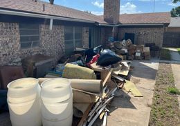 Piles of trash and bulky debris stacked outside a house along a walkway and brick wall.