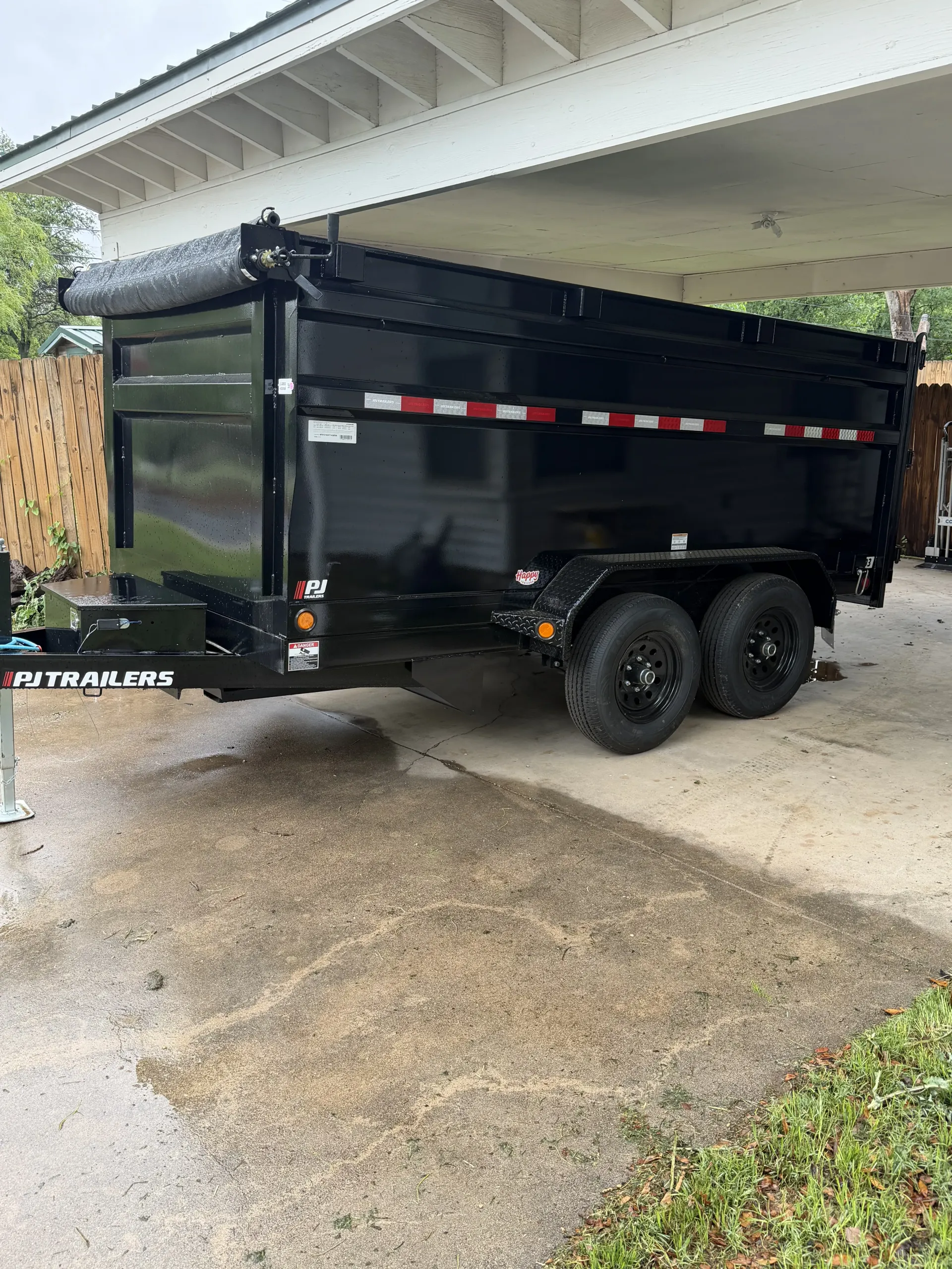 Black dump trailer parked under a carport on a gravel driveway.