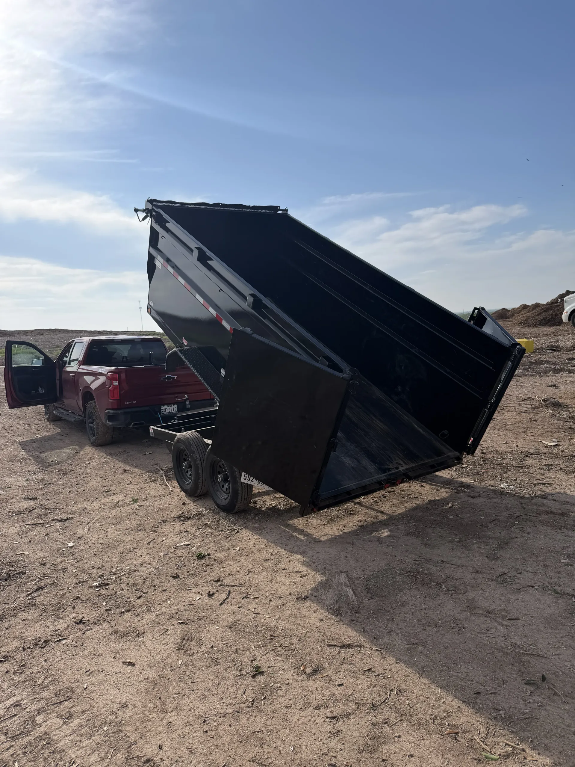 Black dump trailer raised at an angle in a gravel lot beside a red SUV under a blue sky.