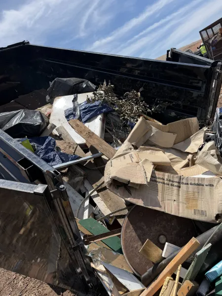 Broken furniture and cardboard piled in a dumpster under a blue sky.