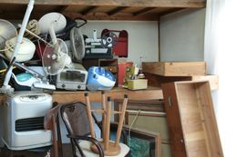 Cluttered storage room with stacked appliances, furniture, and wooden boards under shelves.