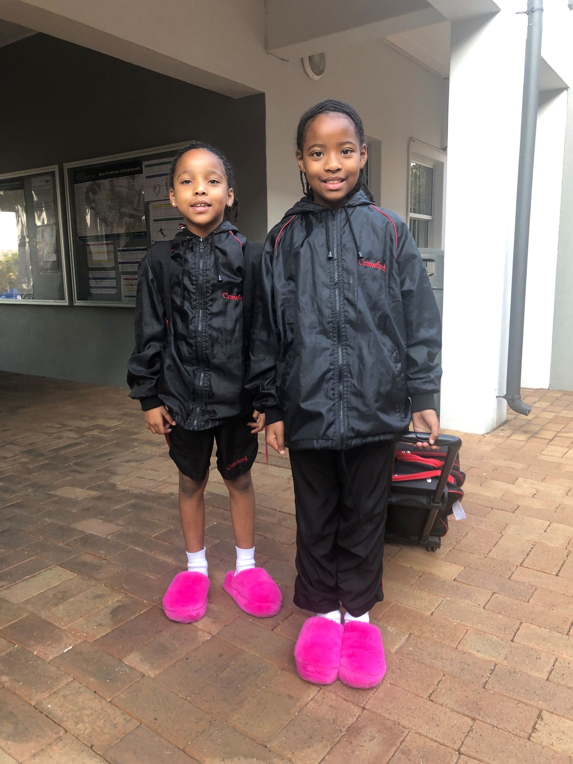 Two young girls wearing pink slippers and black jackets are standing next to each other.