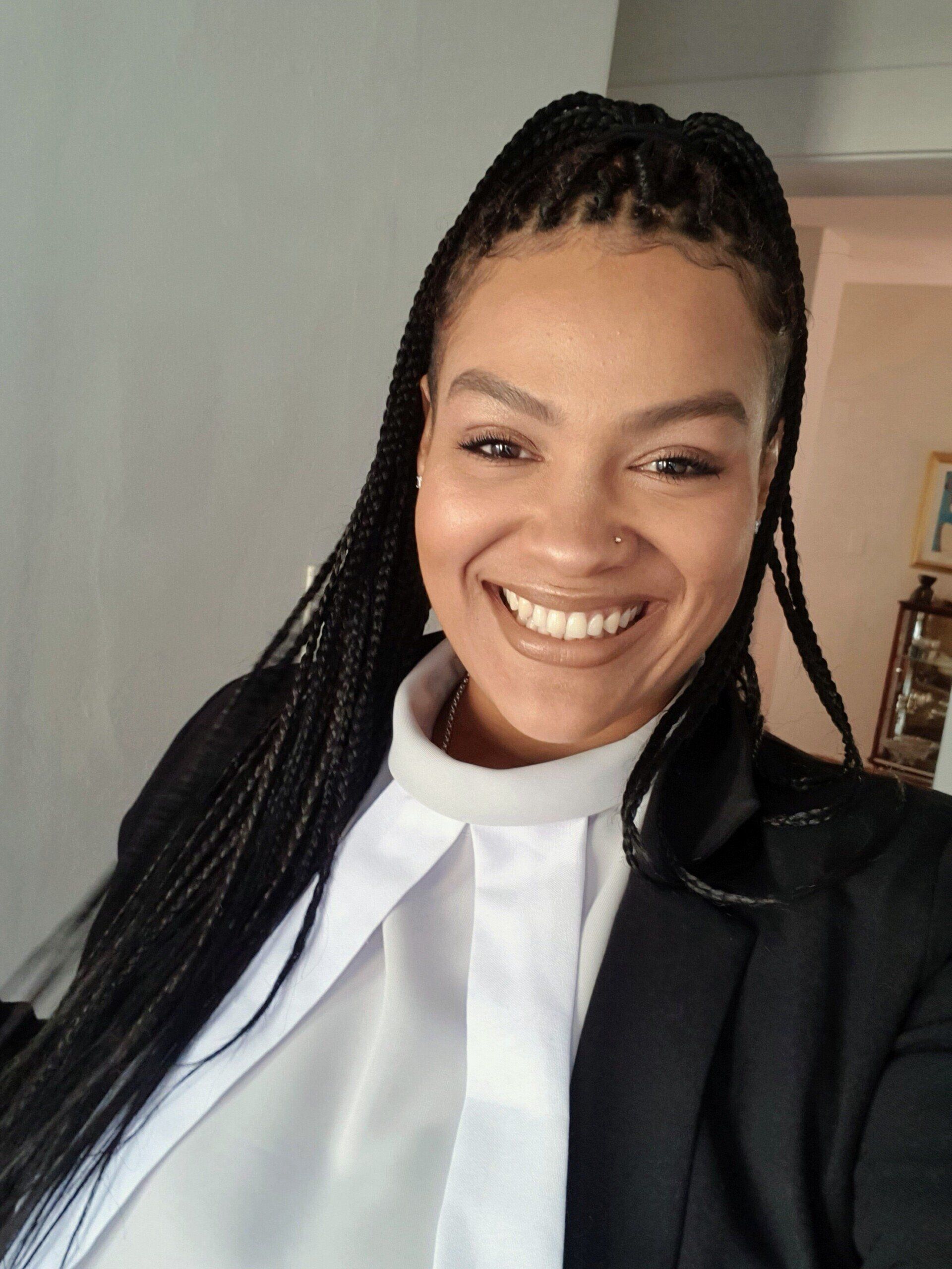 A woman with braids is smiling and wearing a white shirt and a black jacket.