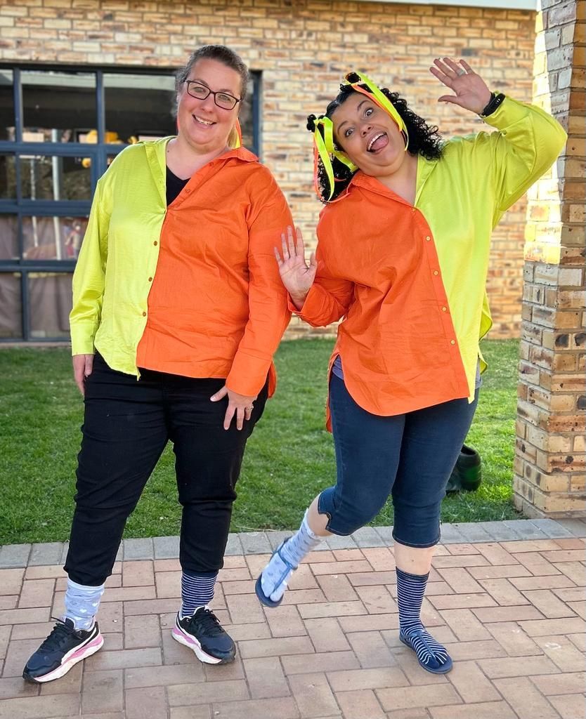 Two women wearing orange and yellow shirts are standing next to each other on a sidewalk.
