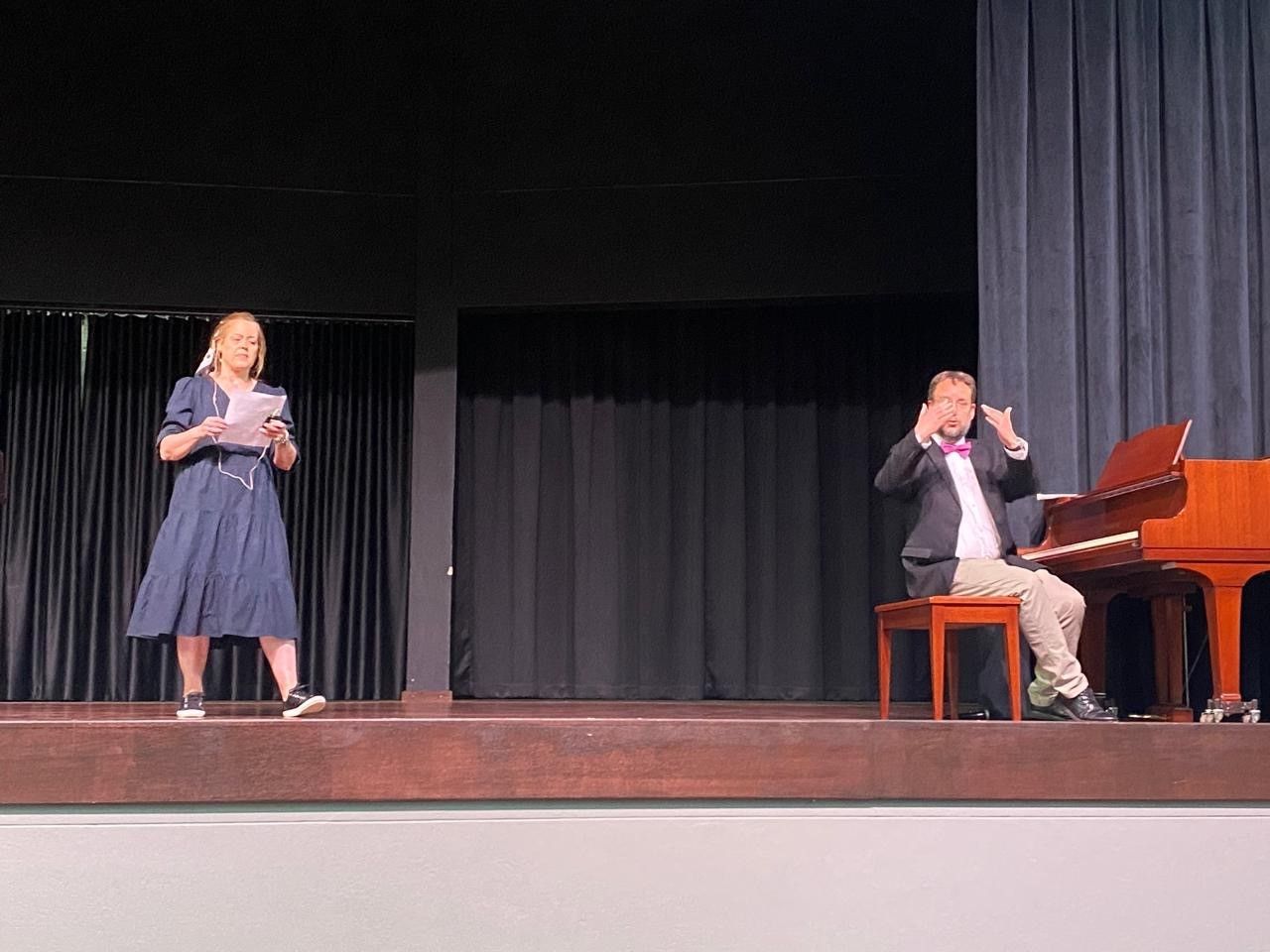 A woman is standing on a stage next to a man sitting at a piano.