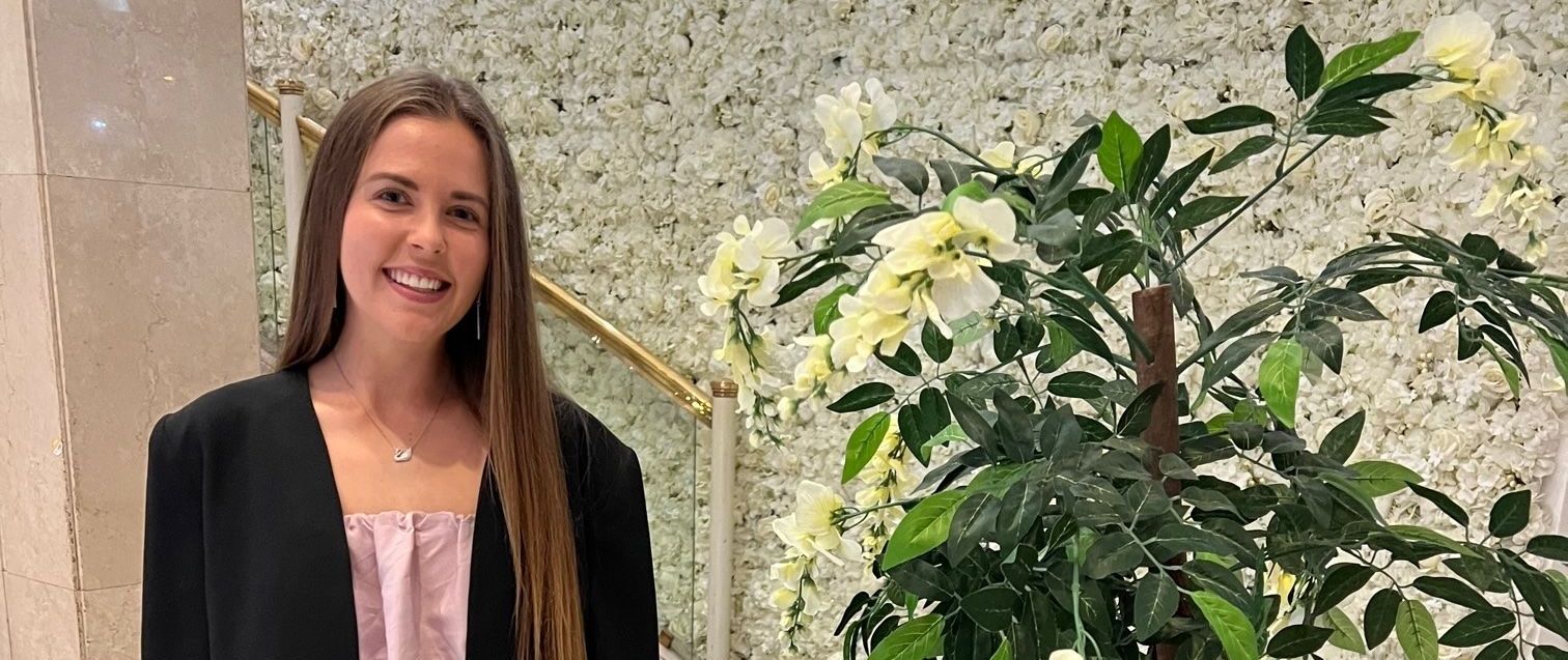 A woman in a black jacket is standing in front of a flower wall.