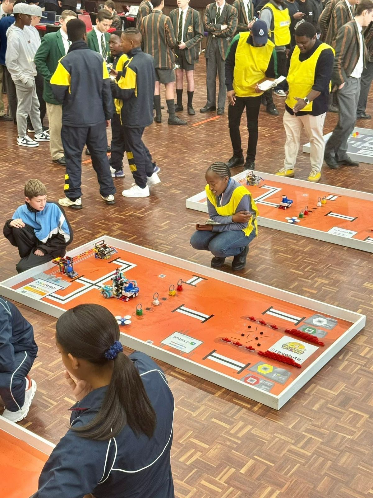 Students and monitors with robots on orange game boards in a competition.