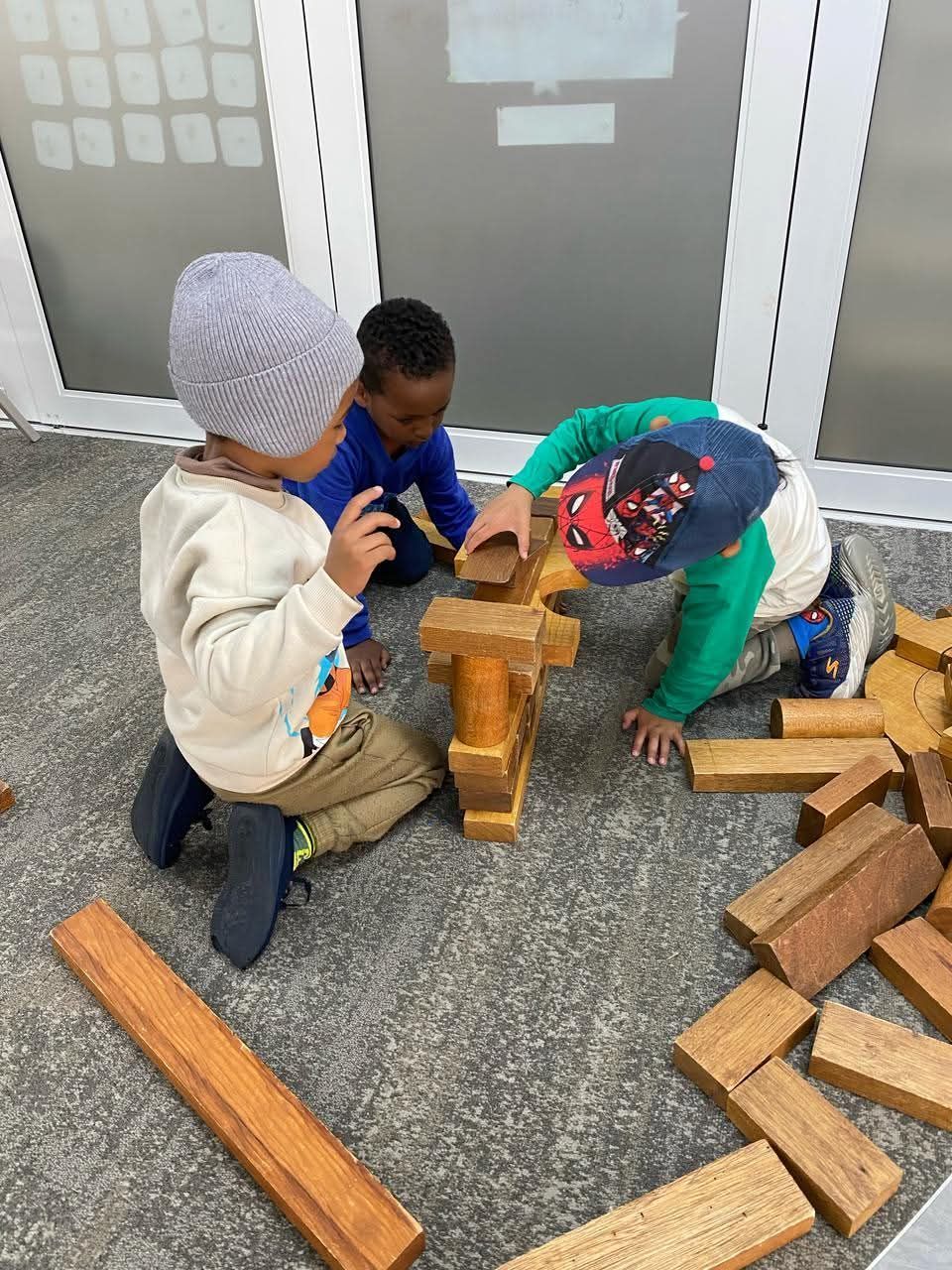 Three children building a structure with wooden blocks on a carpeted floor.