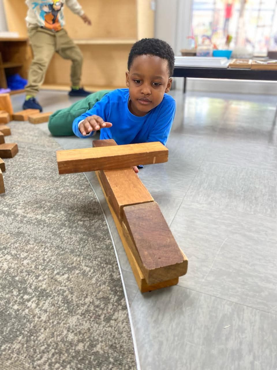 Boy building with wooden blocks on a floor, focused, other child in the background.