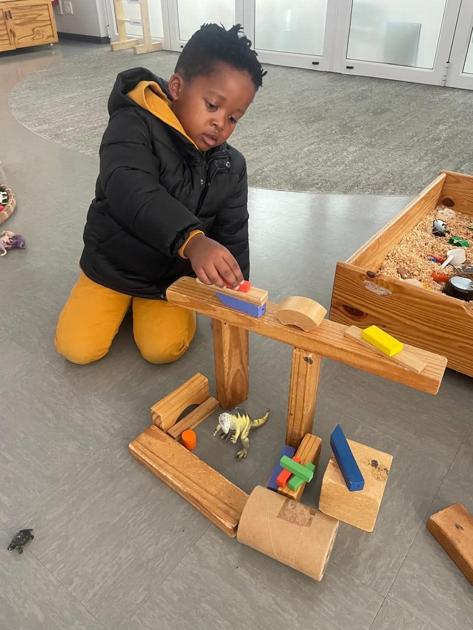 Child building with wooden blocks and small toys. Indoors, wearing a black jacket and yellow pants.