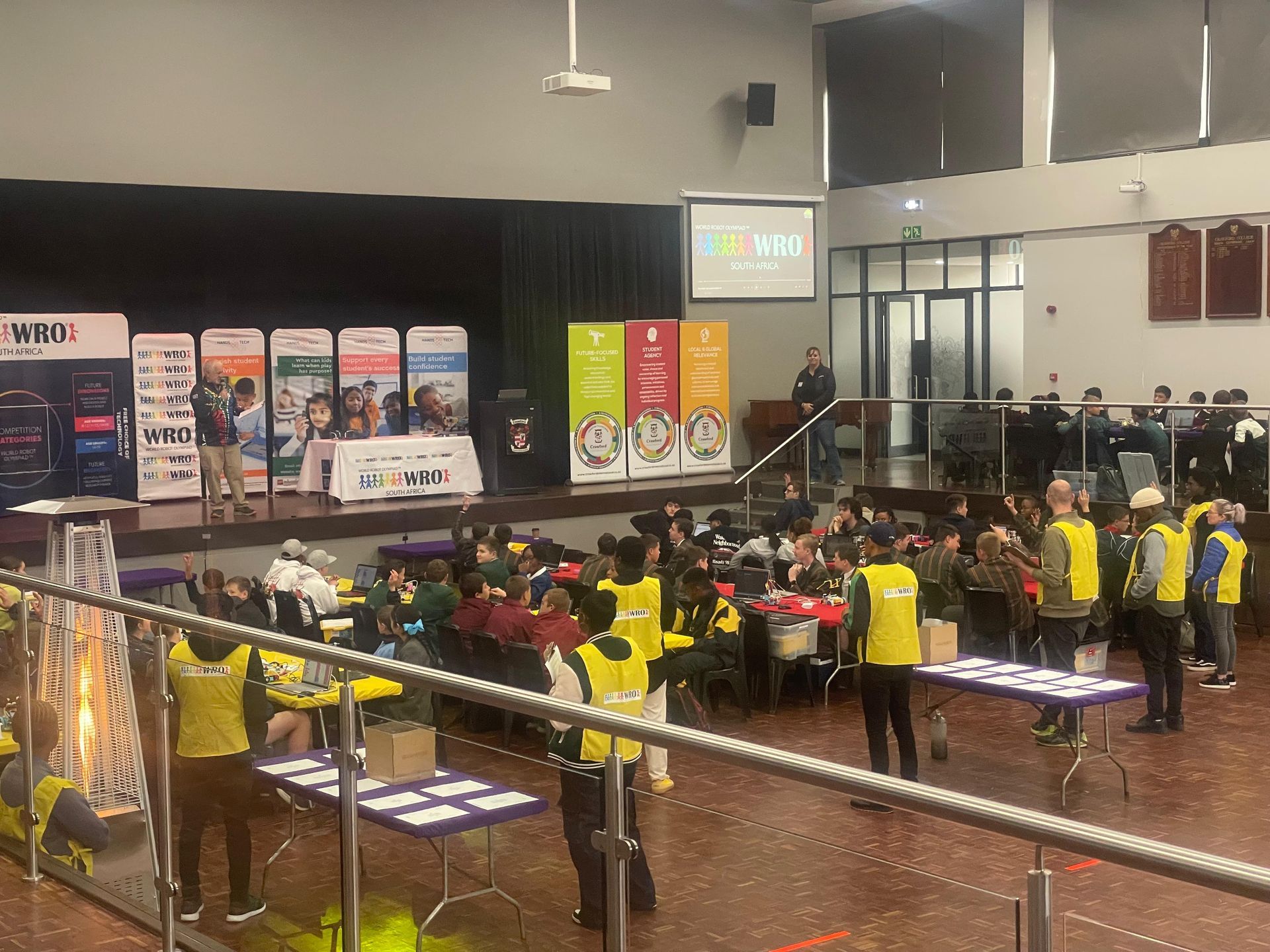 People at an event with a presenter on stage. Yellow vests, banners, and tables are present in a hall.