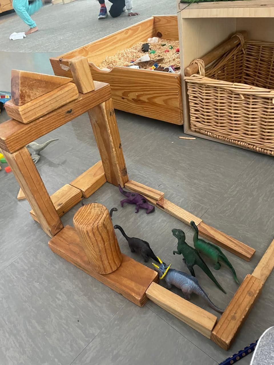 Wooden blocks built as a dinosaur enclosure with toy dinosaurs inside; a child plays.