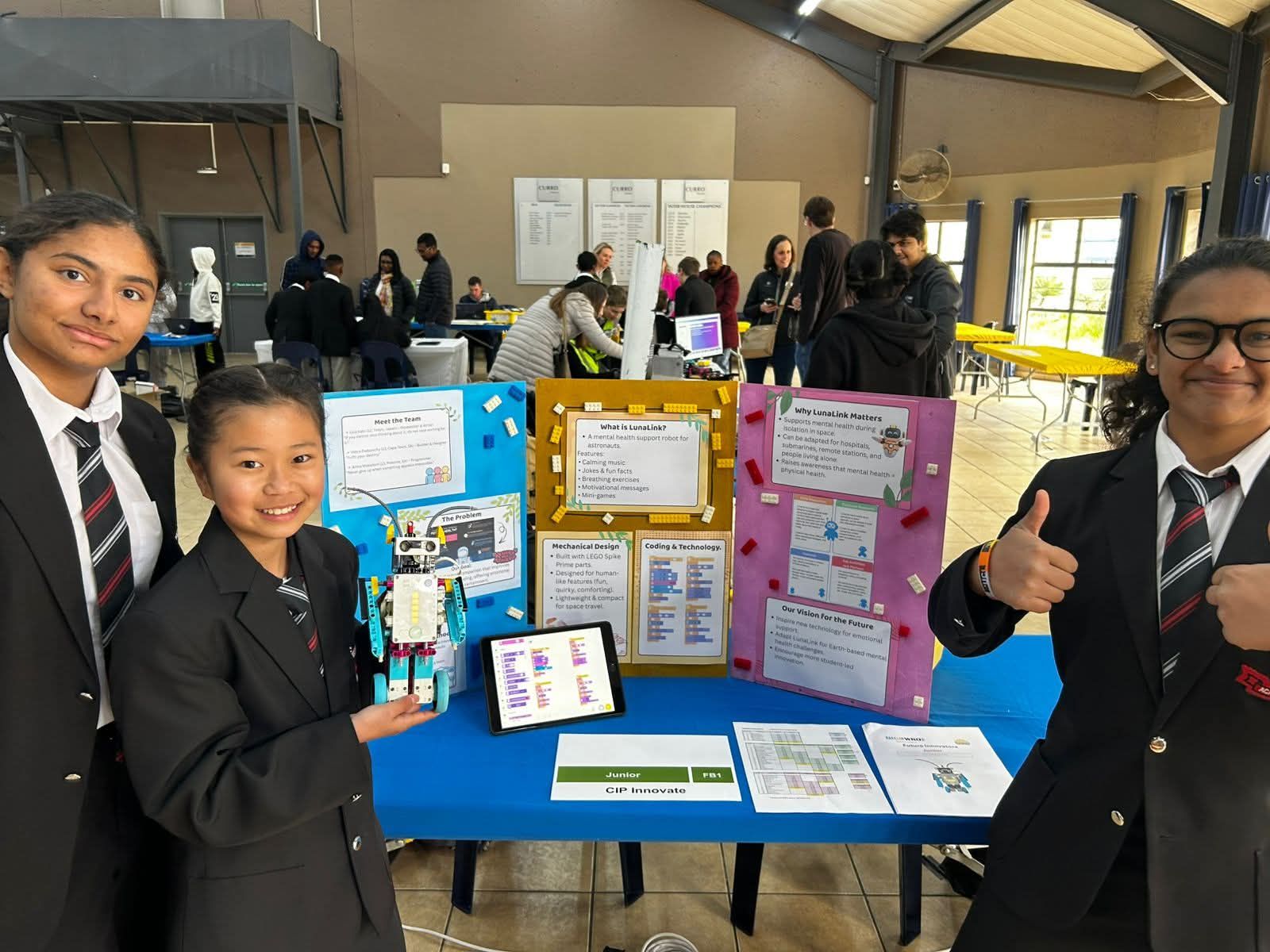 Three students present a project at a science fair; they stand with colorful posters and a robot on a blue table.