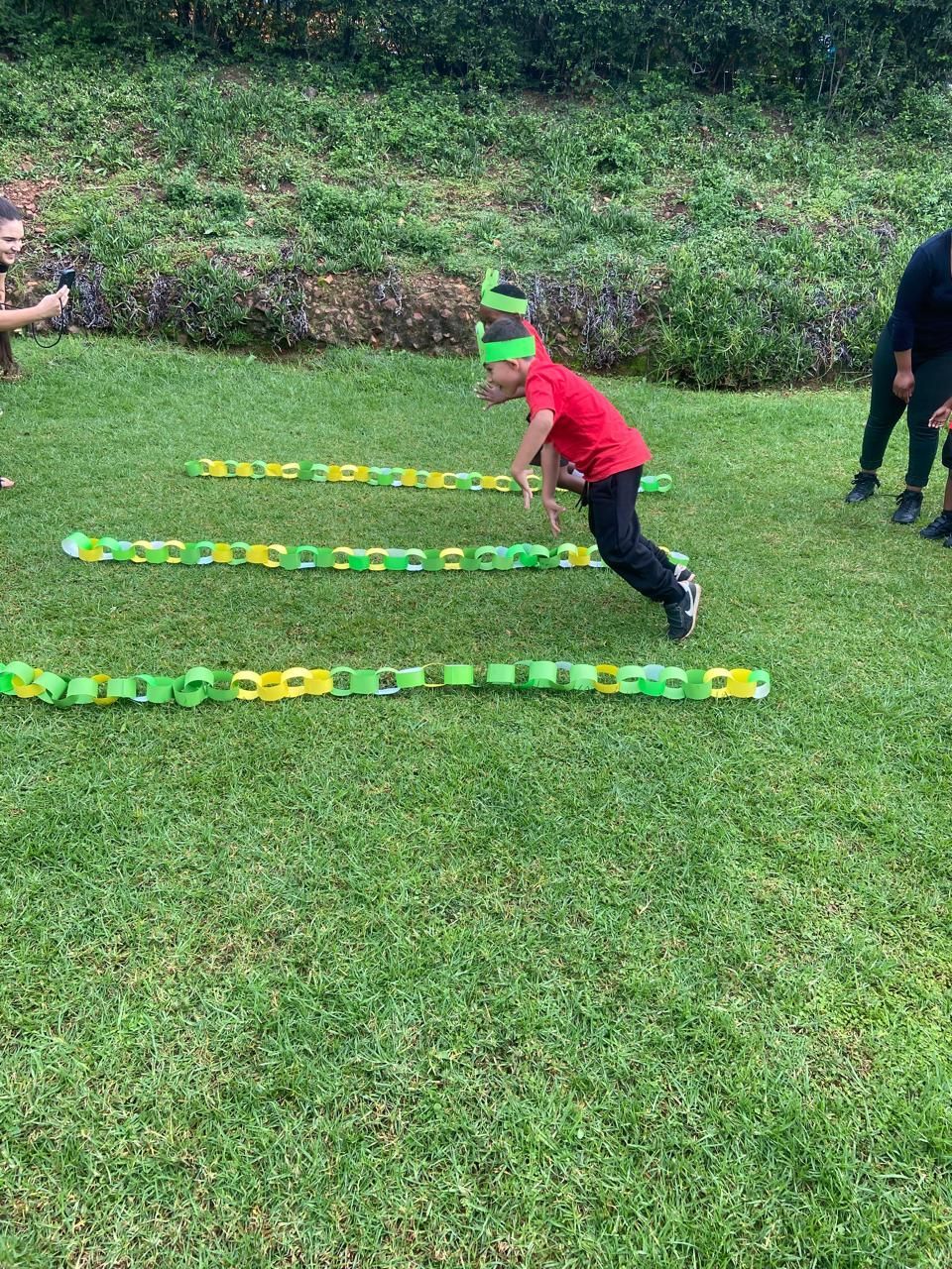 Child in red shirt jumps over green and yellow obstacles on grass.