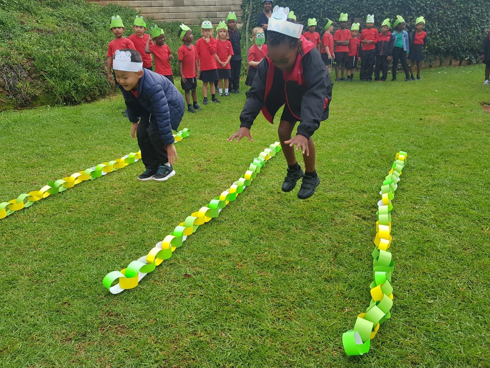 Children jumping over colorful paper chains on grass, other kids watching.