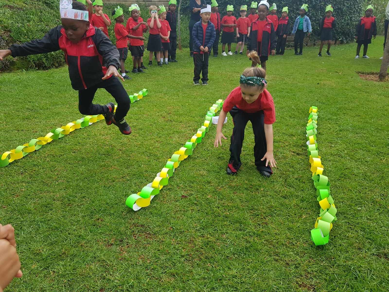 Children jumping over paper chain obstacles on grass. Other kids watch in background.