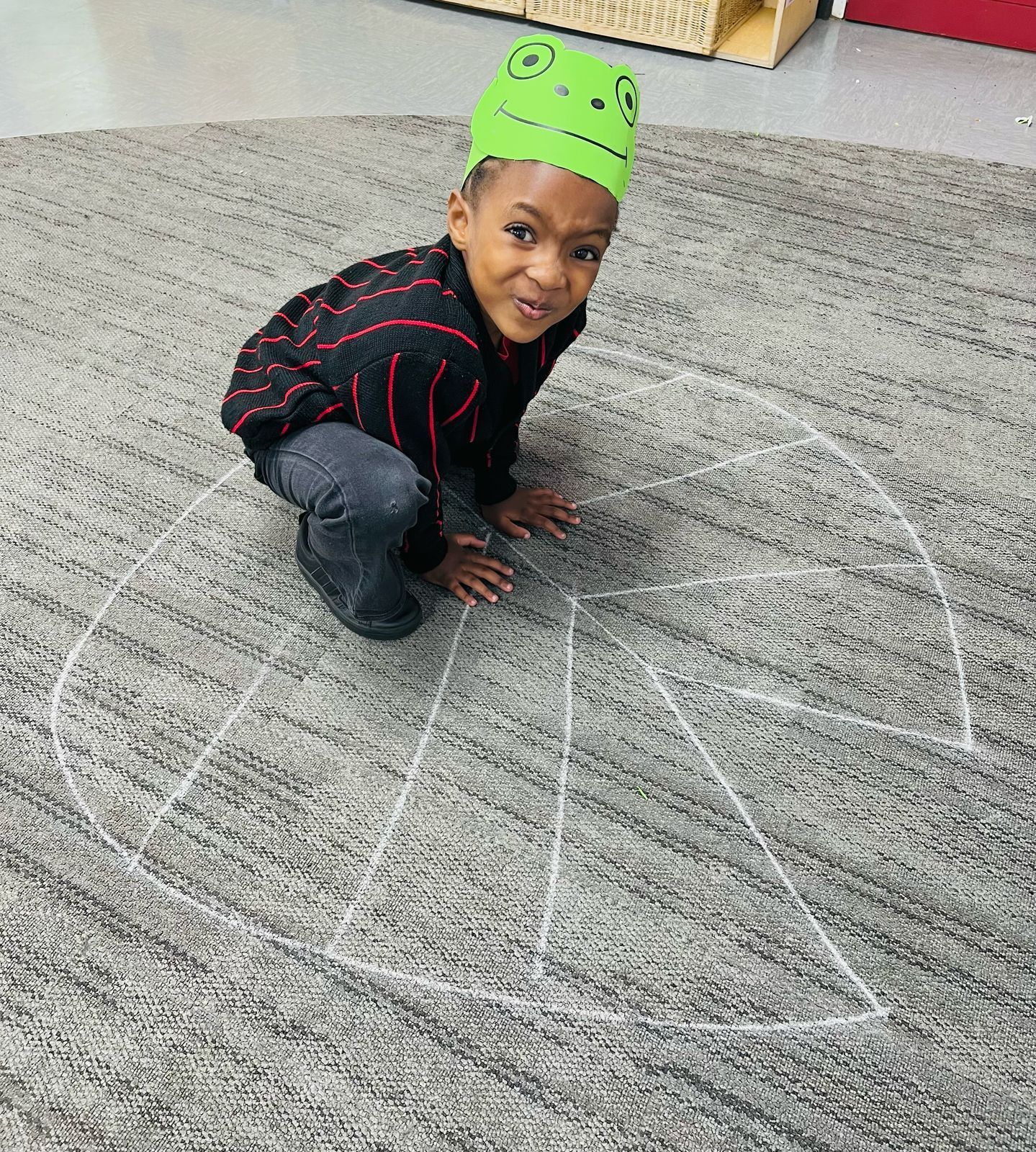 Young child wearing a frog hat kneels on a carpet with a chalk-drawn pie chart and smiles.