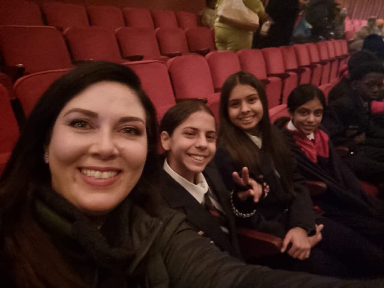A woman smiles, taking a selfie with three children in red theater seats.