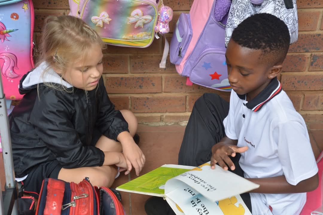 Two students reading a book together near backpacks, one child is pointing at the book.