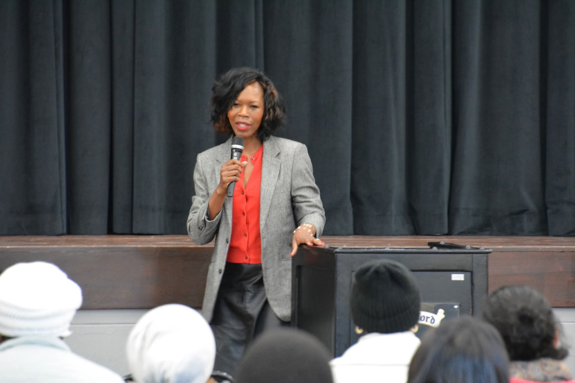 Woman speaking into a microphone on stage, wearing a red top and gray jacket; dark curtain backdrop.