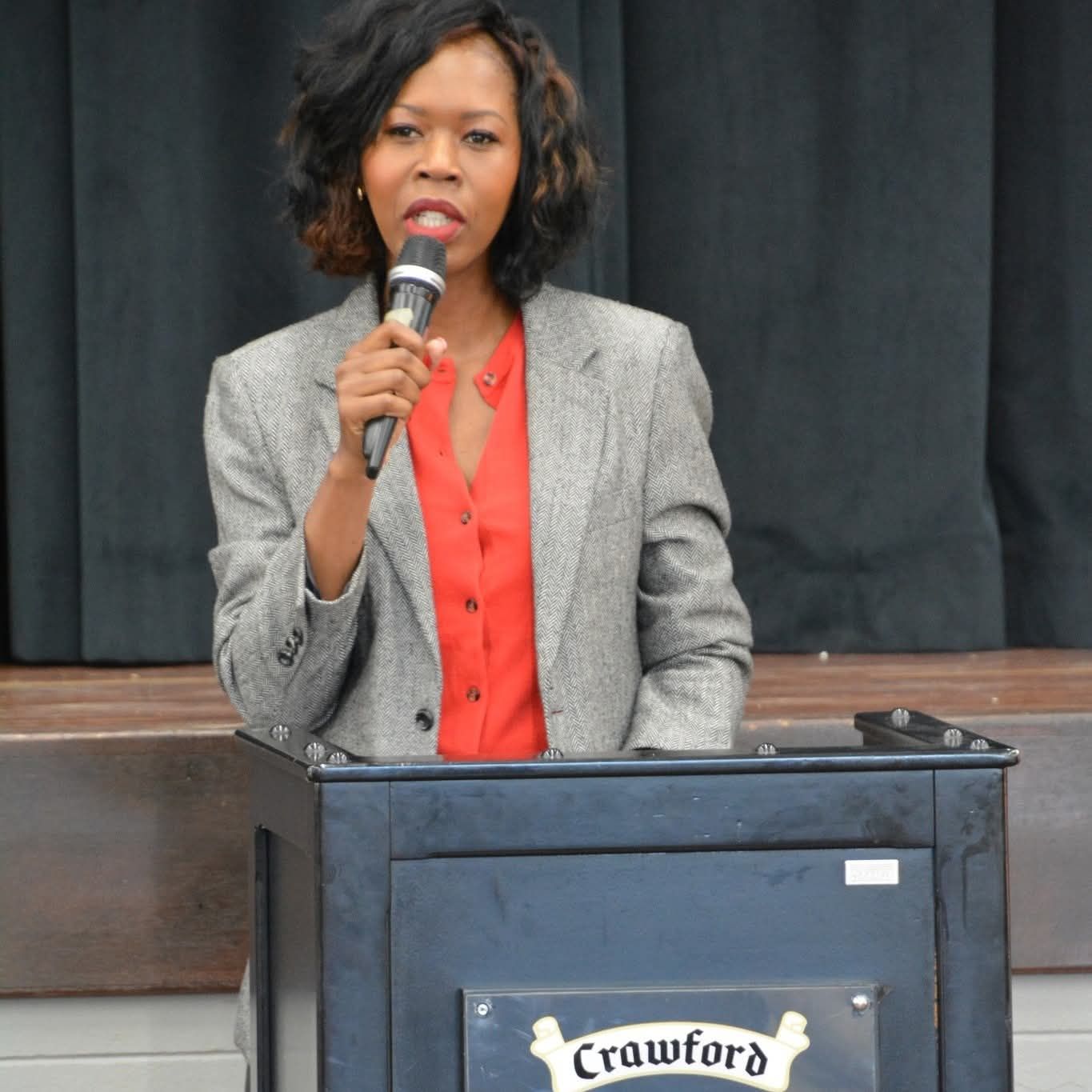 Woman speaking at a podium, wearing a gray blazer and red shirt, in a room with a stage.